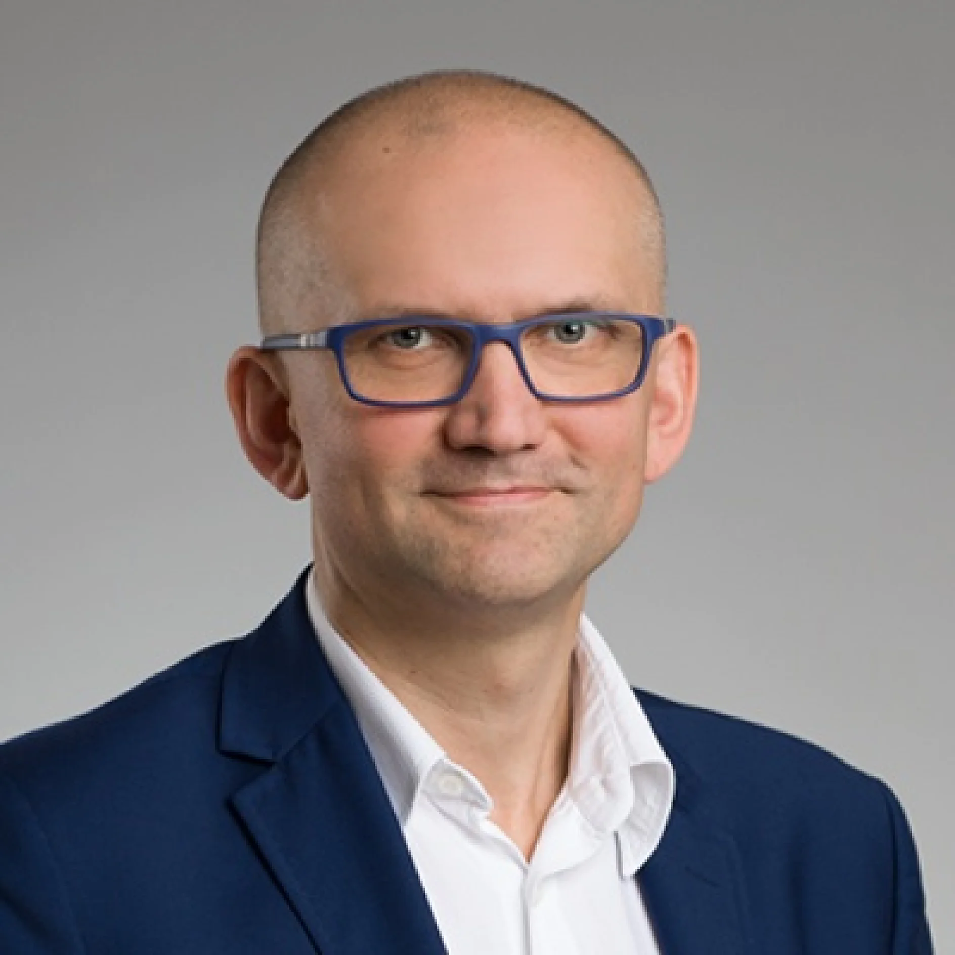 Osoba nosząca okulary jest ubrana w granatowy garnitur i białą koszulę. Tło jest w jednolitym szarym kolorze. (This text has been generated by AI)
