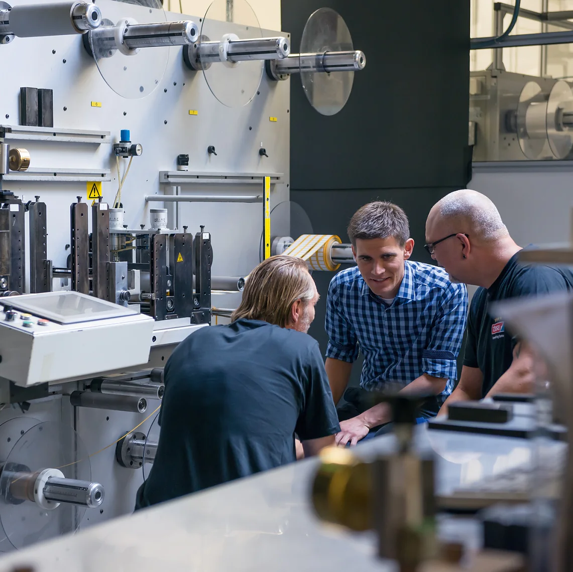Three men are gathered around a large industrial machine in a factory setting. They appear to be engaged in discussion. The machine has various levers and components, and there are rolls of tesa tape in the background. (This text has been generated by AI)