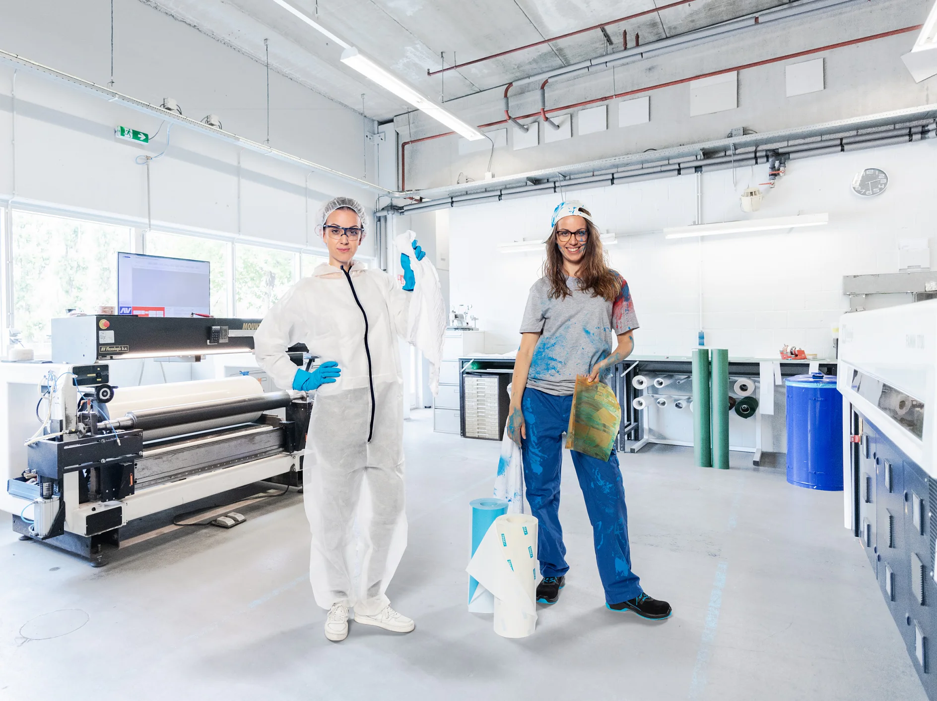 Two people stand in a printing workshop. One wears a white protective suit and goggles, holding a roll of tesa tape. The other is dressed in casual clothes with a cap, holding a large print. The background features printing equipment and rolls of material. (This text has been generated by AI)