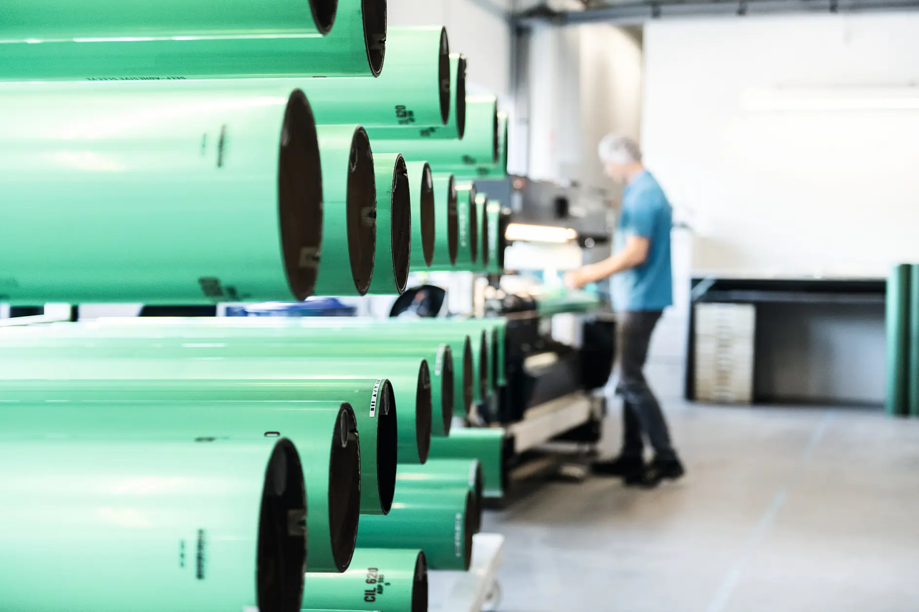 Rows of large green cylindrical tubes are lined up in a workshop. A person wearing a blue shirt is blurred in the background, working with tesa tape and other equipment. The setting appears to be an industrial or manufacturing environment.