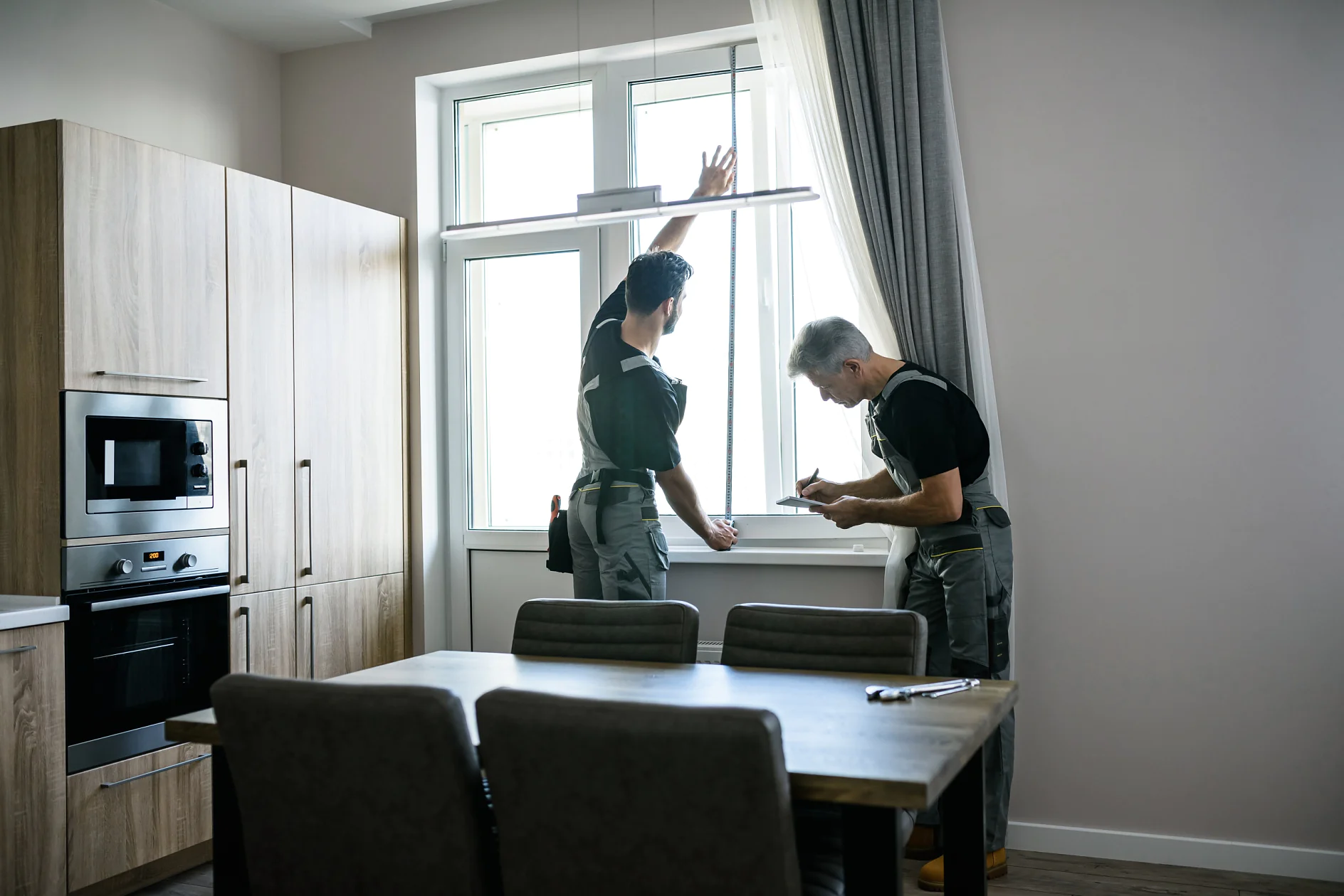 Two workers in overalls are installing or inspecting a window in a modern kitchen. One stands on a ledge, adjusting the window frame with tesa tape, while the other uses a tool or takes notes. A dining table and chairs are in the foreground. (This text has been generated by AI)
