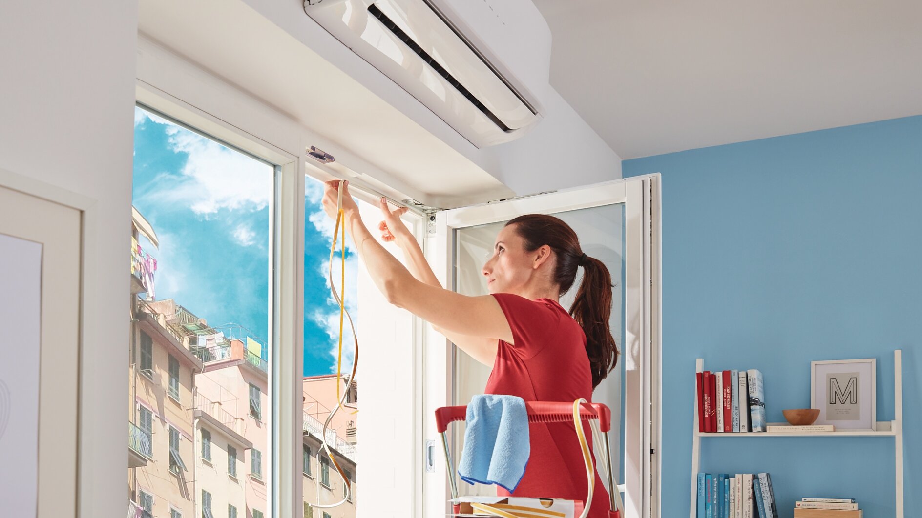Osoba na drabinie instaluje lub naprawia klimatyzację w pobliżu okna w słonecznym pokoju. Przenosi kable, używając taśmy tesa do ich zabezpieczenia, a na drabinie leży ręcznik. W tle widoczne są regały z książkami i dekoracjami. (This text has been generated by AI)
