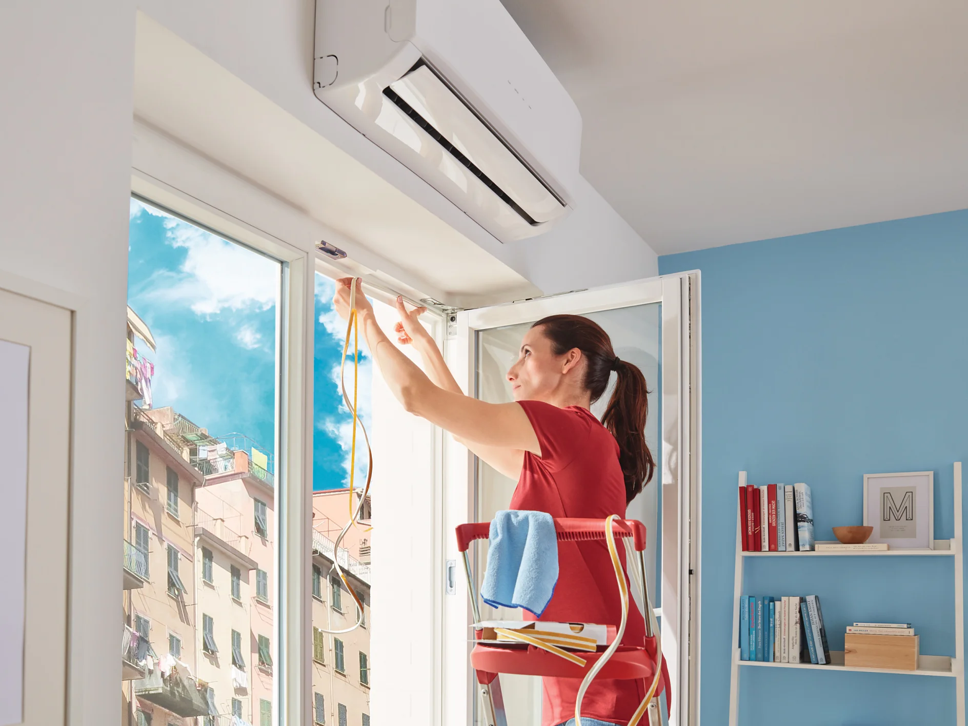 Osoba na drabinie instaluje lub naprawia klimatyzację w pobliżu okna w słonecznym pokoju. Przenosi kable, używając taśmy tesa do ich zabezpieczenia, a na drabinie leży ręcznik. W tle widoczne są regały z książkami i dekoracjami. (This text has been generated by AI)