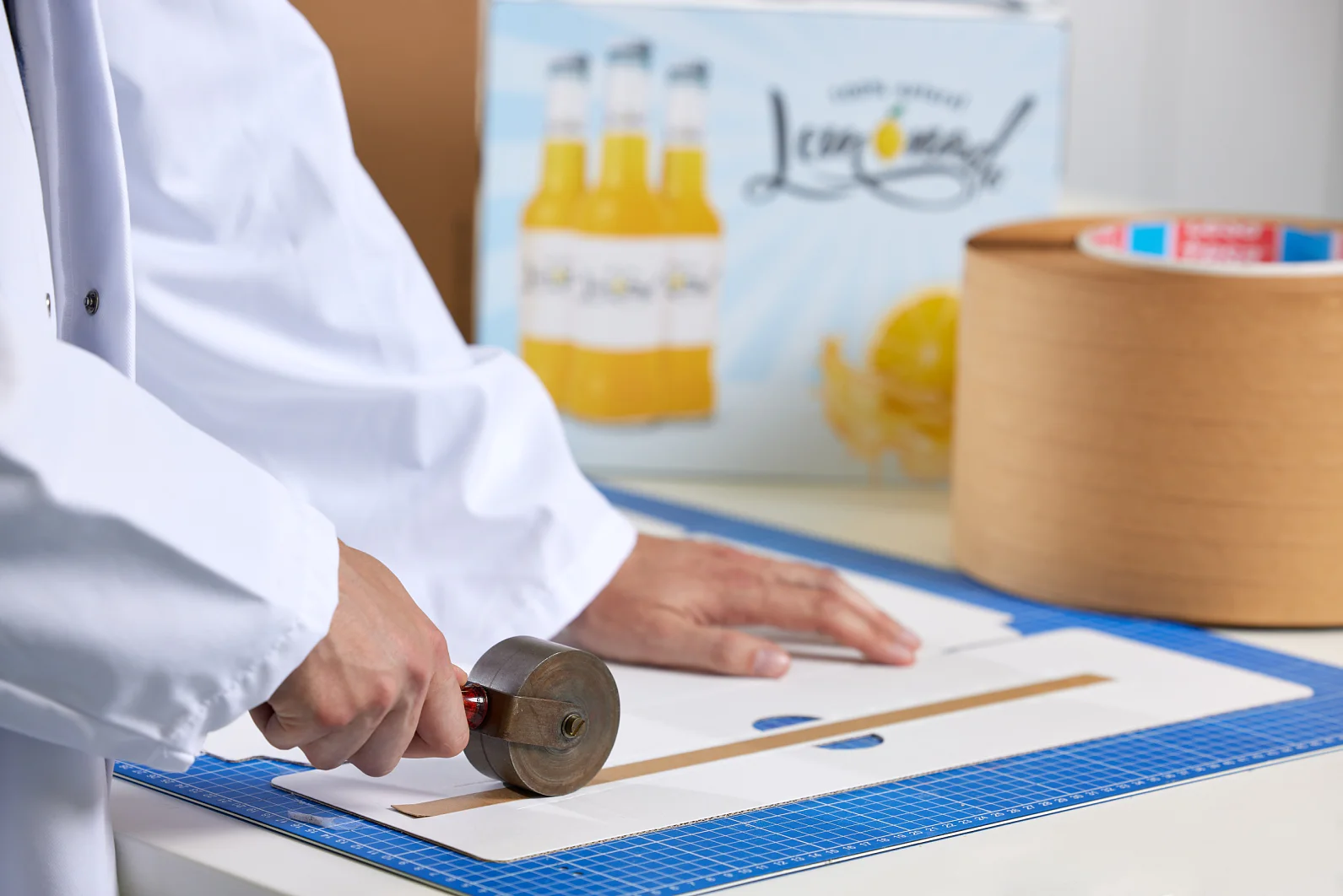 A person in a white coat uses a hand roller on a flat cardboard box on a blue cutting mat. In the background, theres a box of bottled lemonade. A large roll of brown tesa tape is visible on the right. (This text has been generated by AI)