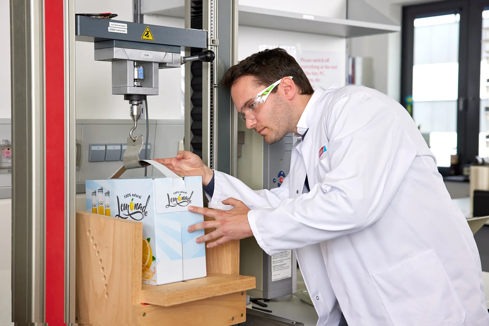 A man in a white lab coat and safety glasses examines a box of juice cartons on a testing machine in a lab. The machine appears to be applying pressure to the box secured with tesa tape. There are notices and equipment visible in the background. (This text has been generated by AI)