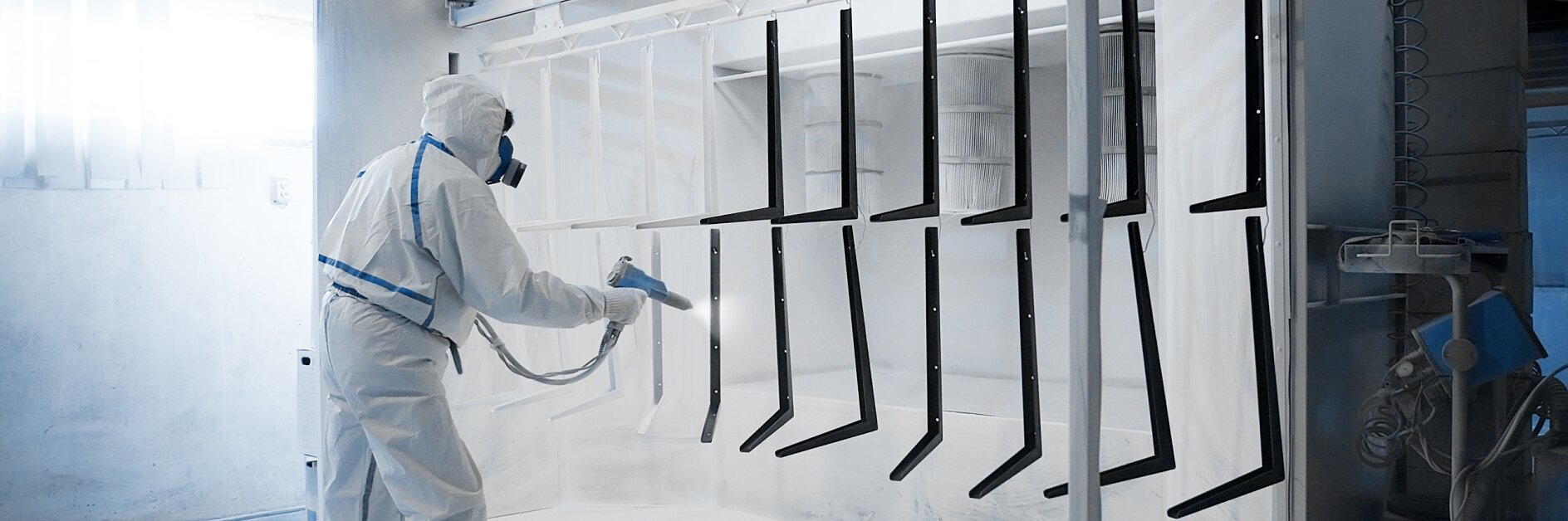 A person wearing protective gear is using a spray gun in an industrial setting. They are applying a coating to metal objects hanging in a spray booth. The environment appears clean and organized, with bright lighting. While working, the person relies on tesa tape for precise masking and protection of certain areas during the spraying process. (This text has been generated by AI)