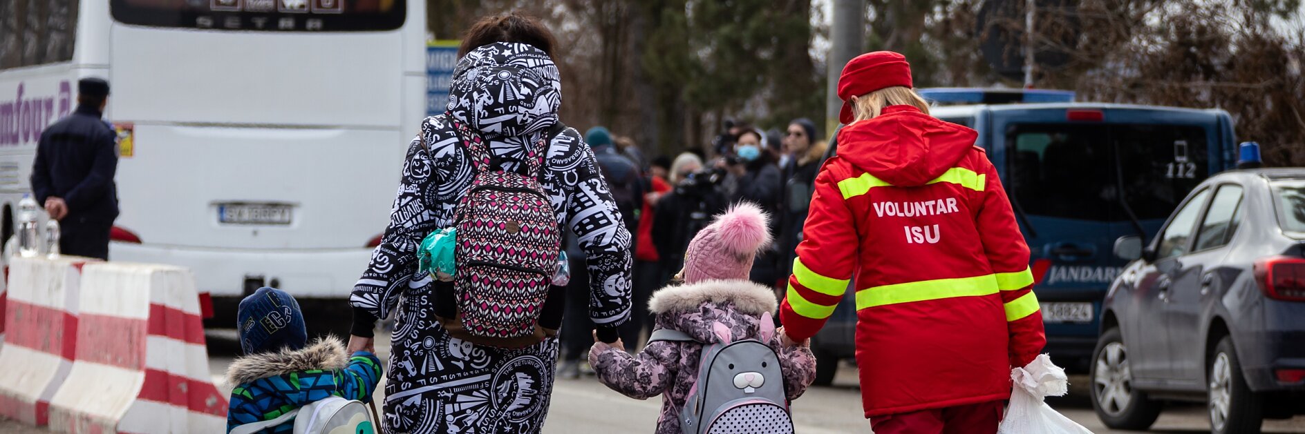 A woman and two children walk down a street near parked vehicles. The children wear backpacks and winter clothing. A volunteer wearing a red uniform assists them, using tesa tape to secure a large white bag. A bus and several people are visible in the background.
