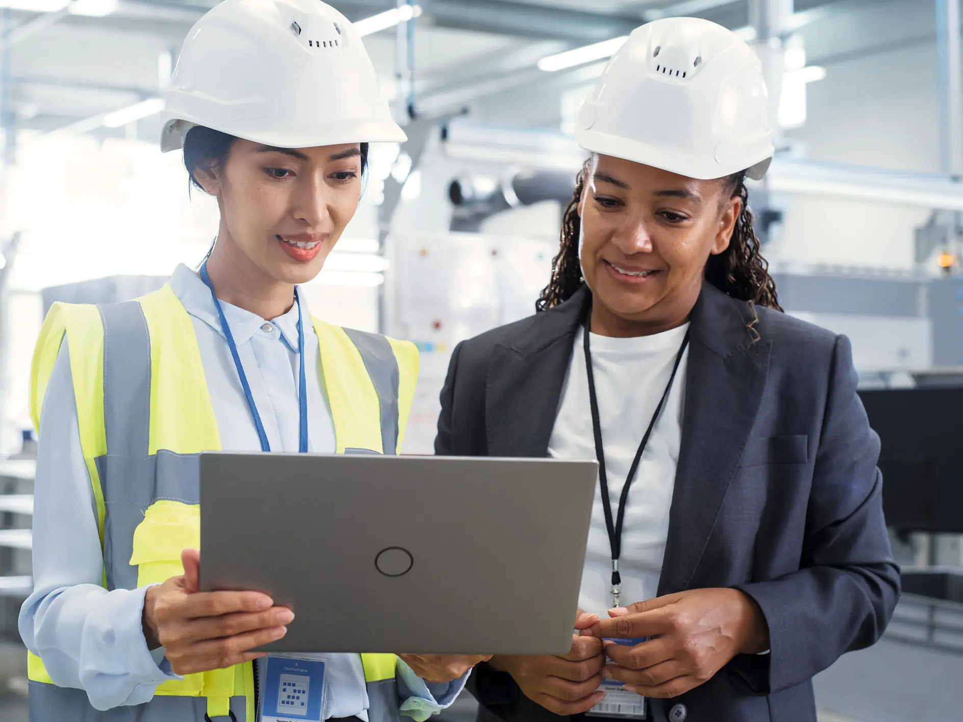 Two professionals in a modern industrial setting looking on a laptop, one wearing a safety vest and hard hat, the other in business attire. (This text has been generated by AI)