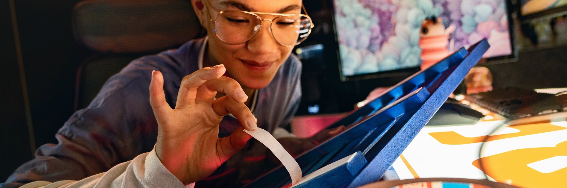 A person with short hair and glasses is sitting at a desk, peeling a strip of tesa tape. The desk is filled with various materials and tools, while a computer monitor in the background shows a colorful image. The setting appears to be creative or craft-oriented.