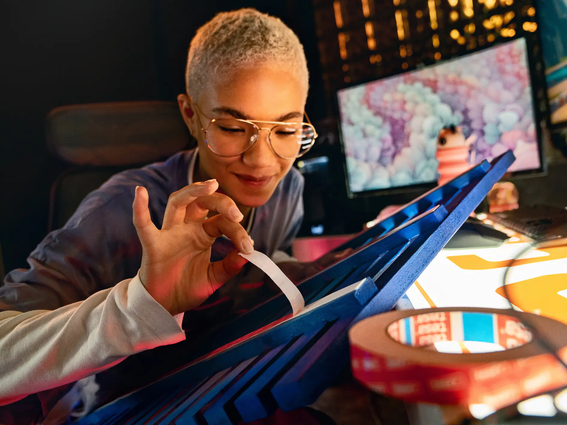A person with short hair and glasses is sitting at a desk, peeling a strip of tesa tape. The desk is filled with various materials and tools, while a computer monitor in the background shows a colorful image. The setting appears to be creative or craft-oriented. (This text has been generated by AI)