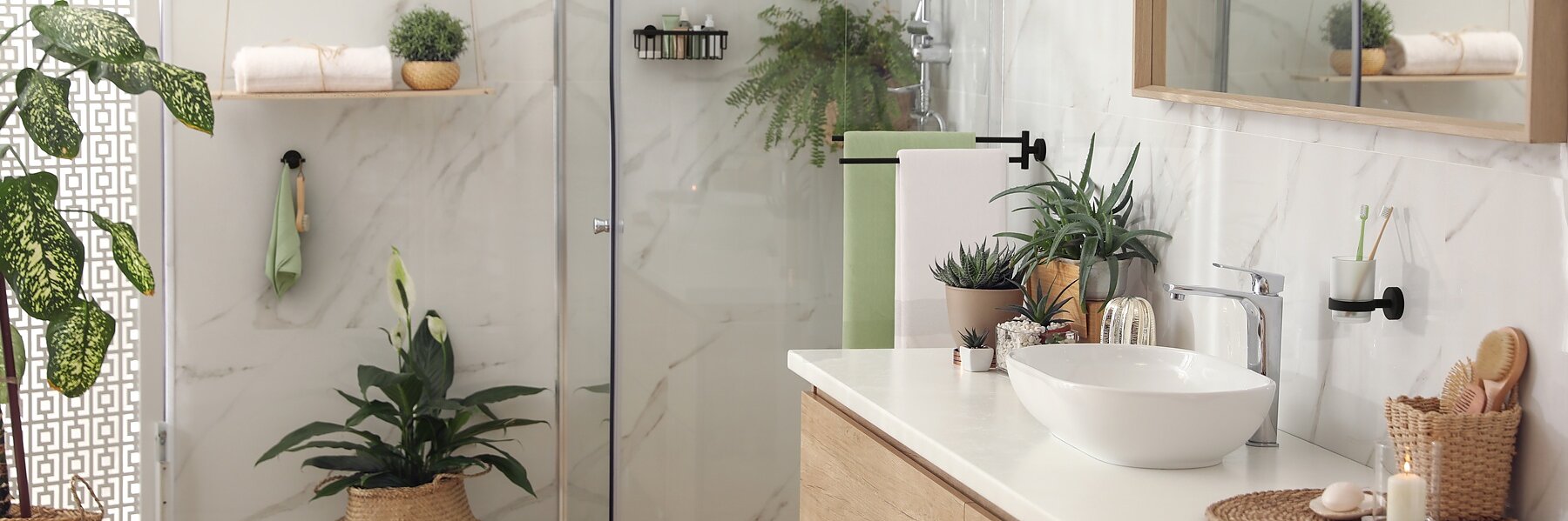 Modern bathroom with a glass shower, wood vanity, and white basin. White marble walls and multiple indoor plants enhance the decor. A mirror hangs above the sink, secured with tesa tape, and there are neatly arranged toiletries on the counter and shelves. (This text has been generated by AI)