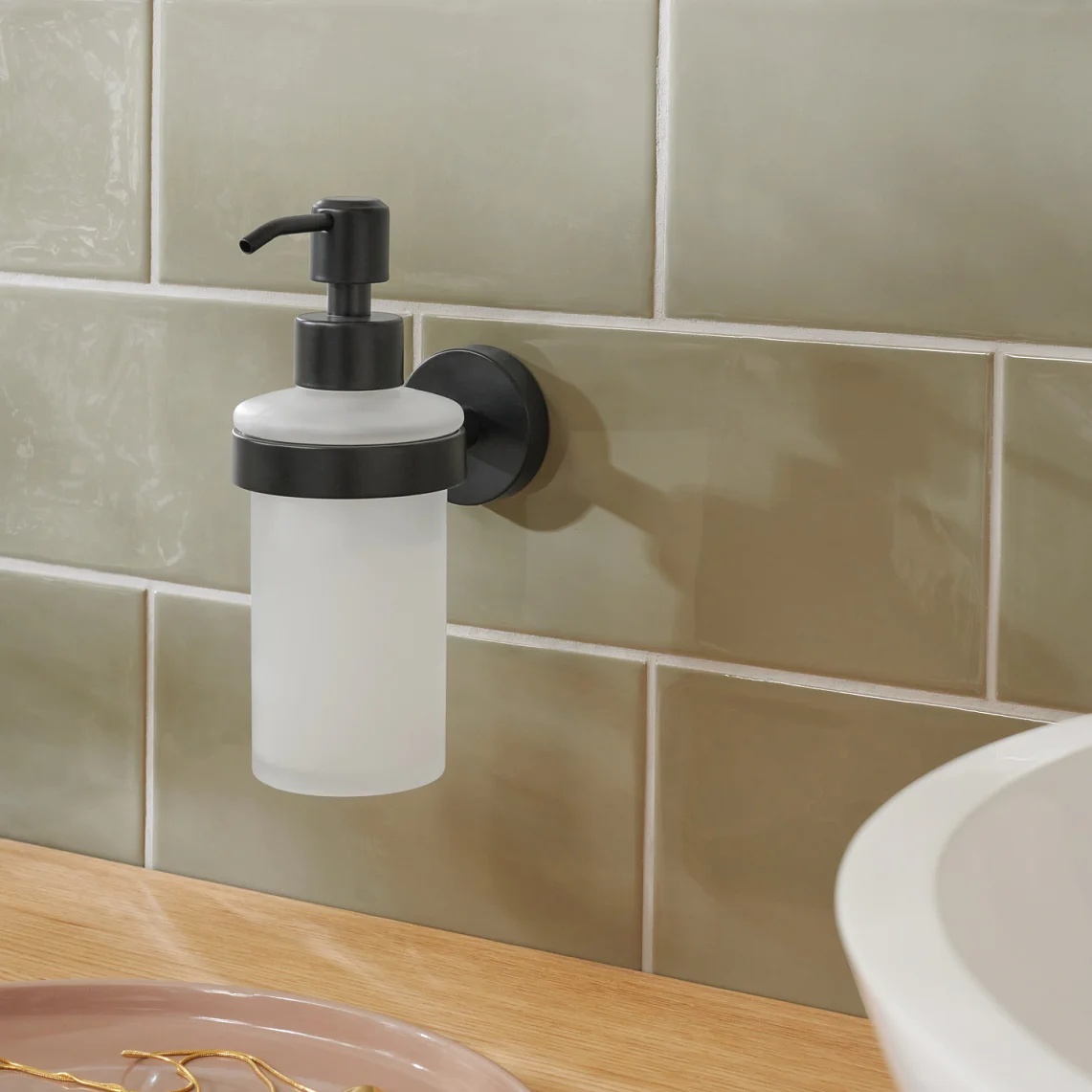 A wall-mounted soap dispenser with a black pump is attached to a tiled wall using tesa tape. The tiles are light green, and a wooden countertop is visible below, partially holding a round white sink and a pink dish with gold jewelry. (This text has been generated by AI)