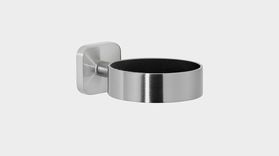 A stainless steel wall-mounted holder with a cylindrical design and a square base. The interior appears to be lined with a dark material. The wall mount is attached to the left side of the holder using tesa tape. (This text has been generated by AI)