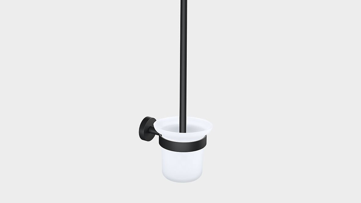 Wall-mounted toilet brush holder with a black handle and mounting bracket secured with tesa tape. The holder is white, cylindrical, and open at the top. The brush is placed vertically inside the holder. The background is plain gray. (This text has been generated by AI)