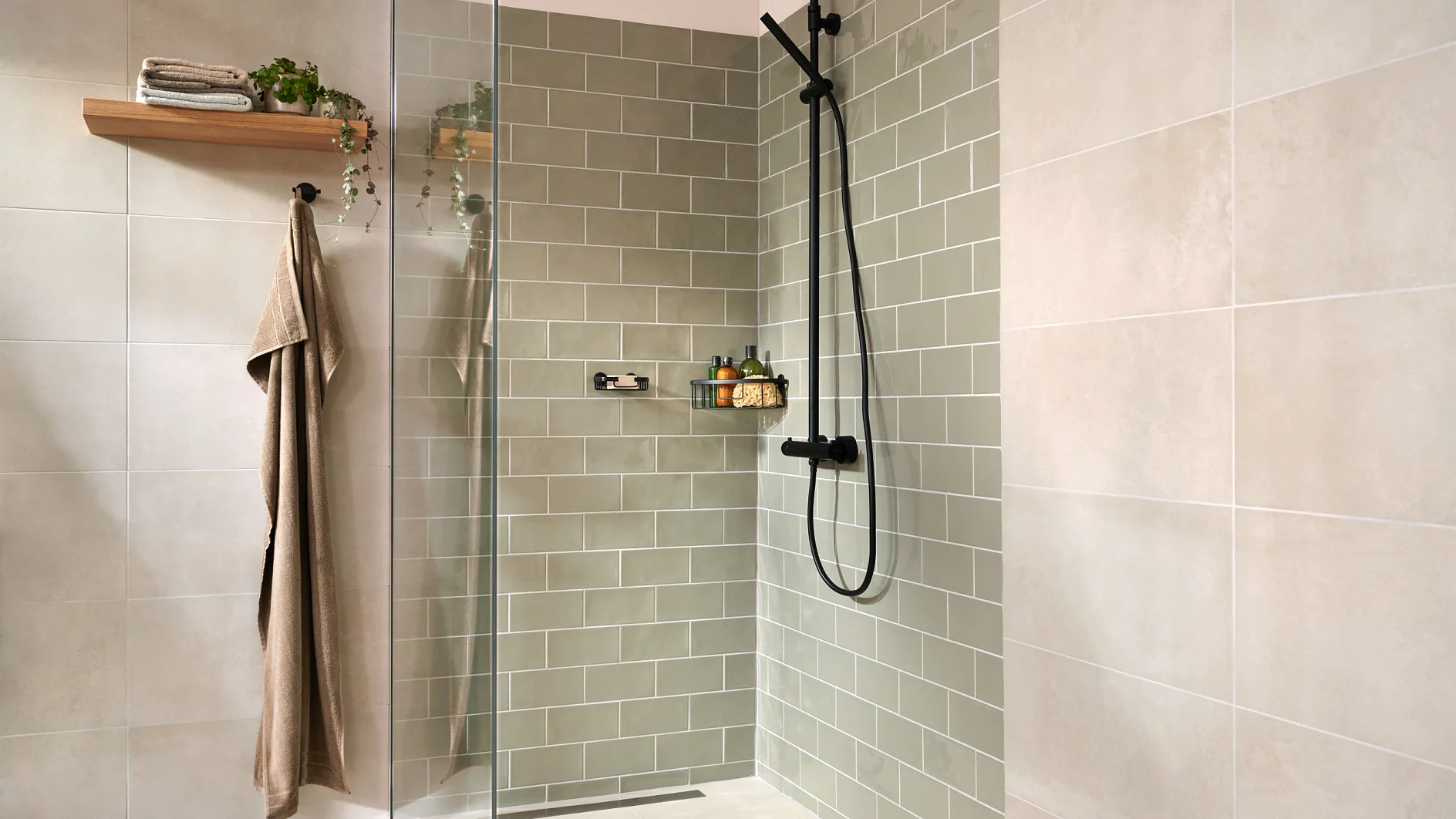 A modern bathroom shower with light green subway tiles and beige wall tiles. It features a glass door, a black shower fixture, a shelf with toiletries, a wooden shelf with plants and towels, secured using tesa tape, and a hanging brown towel. (This text has been generated by AI)