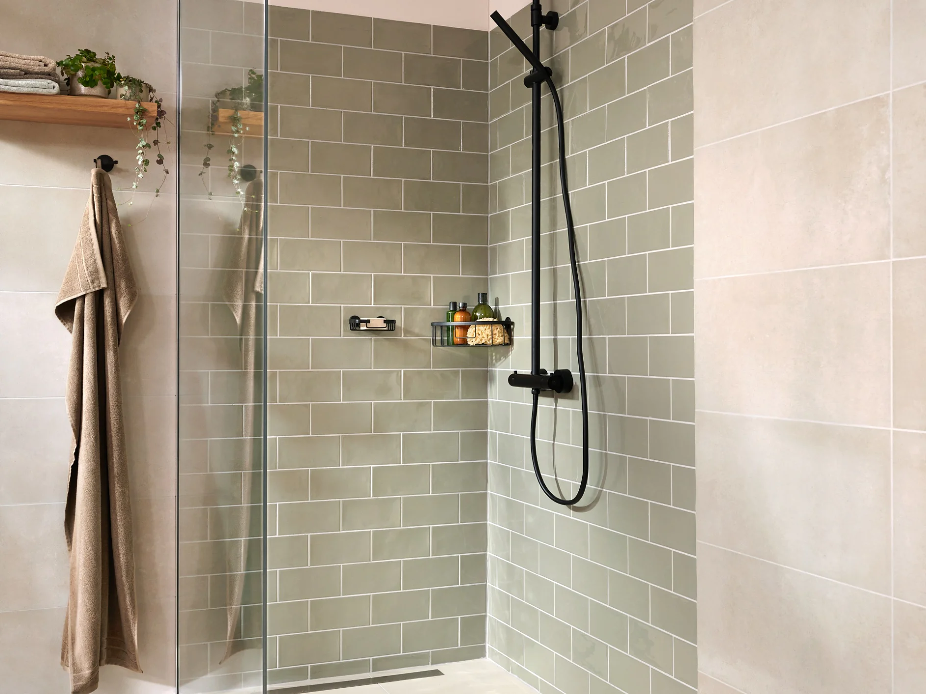A modern bathroom shower with light green subway tiles and beige wall tiles. It features a glass door, a black shower fixture, a shelf with toiletries, a wooden shelf with plants and towels, secured using tesa tape, and a hanging brown towel. (This text has been generated by AI)