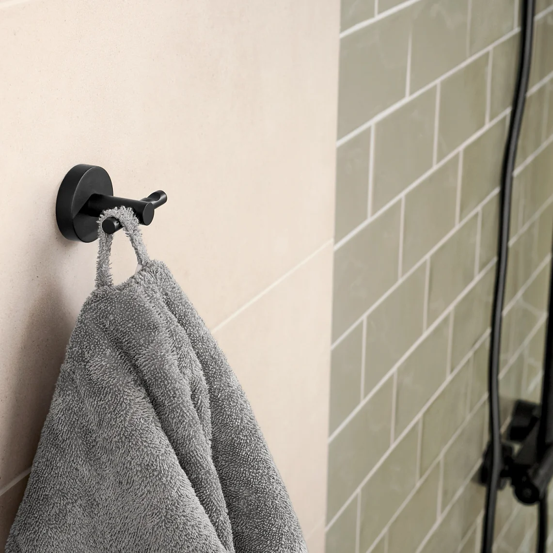 A gray towel hangs on a black hook attached to a beige tiled wall. To the right, there is a partially visible black-framed glass shower door with light green subway tiles. (This text has been generated by AI)