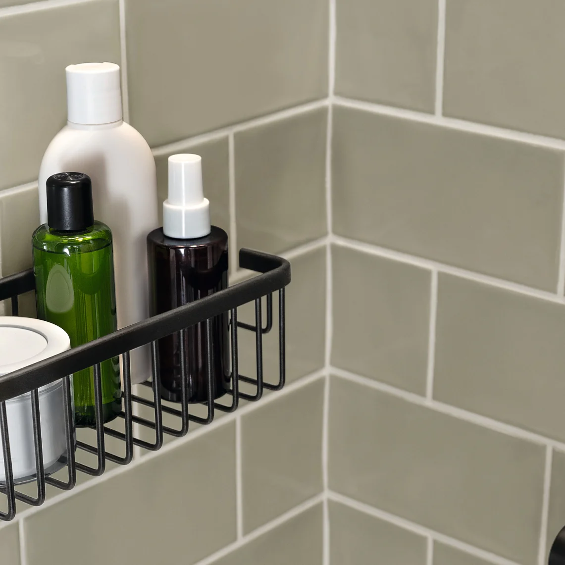 A metal shower caddy is mounted on a light gray tiled wall, holding four bottles of various sizes and shapes. The caddy is fixed in a corner using tesa tape, securely supporting the shampoo or lotion containers. (This text has been generated by AI)