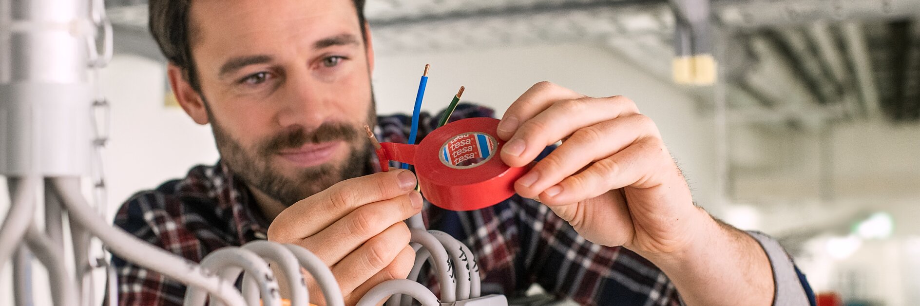 Mężczyzna w kraciastej koszuli używa czerwonego testera elektrycznego do pomiaru wiązki przewodów przymocowanych taśmą tesa do metalowego korytka kablowego w budynku przemysłowym lub handlowym. (This text has been generated by AI)