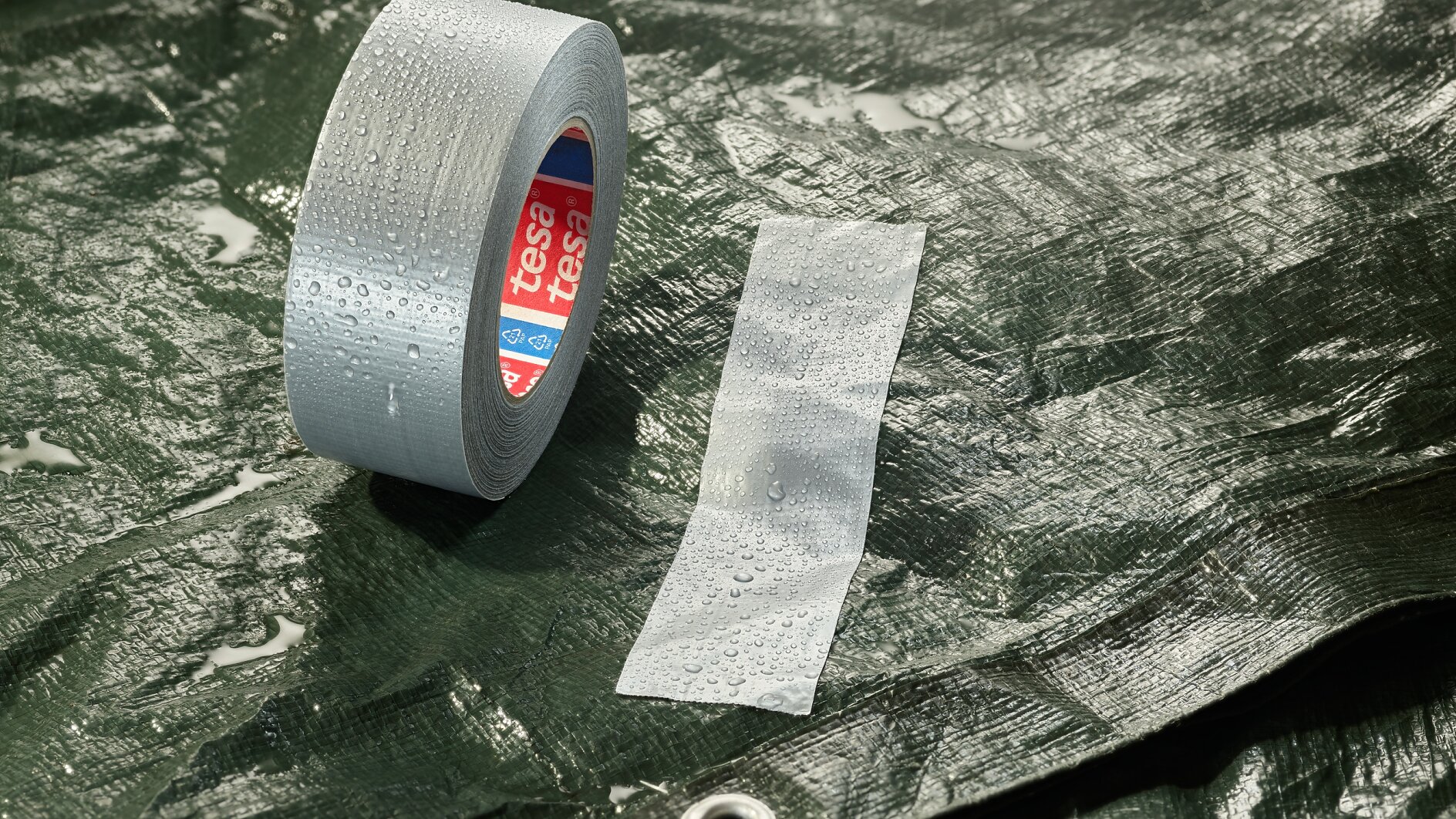 A roll of grey adhesive tesa tape and a strip of tesa tape are placed on a wet, dark green tarpaulin. The tarpaulin has a metal eyelet, and water droplets are visible on the surface. (This text has been generated by AI)