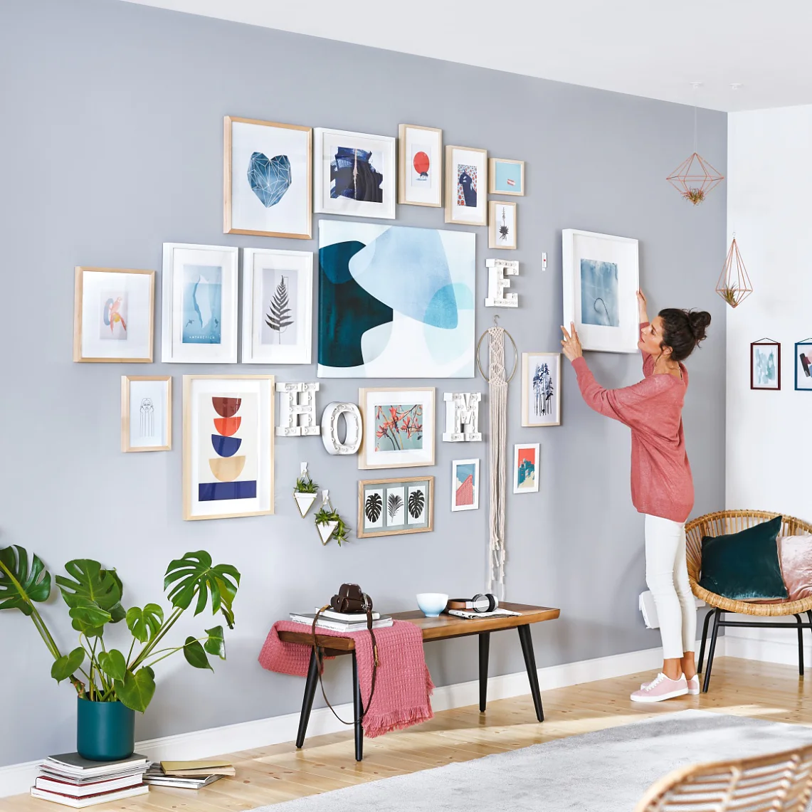 A woman decorates a living room wall with various framed artworks, using tesa tape. The room features a light gray wall, wooden flooring, and modern furniture, including a wooden table and chairs. A kitchen is visible in the background. (This text has been generated by AI)