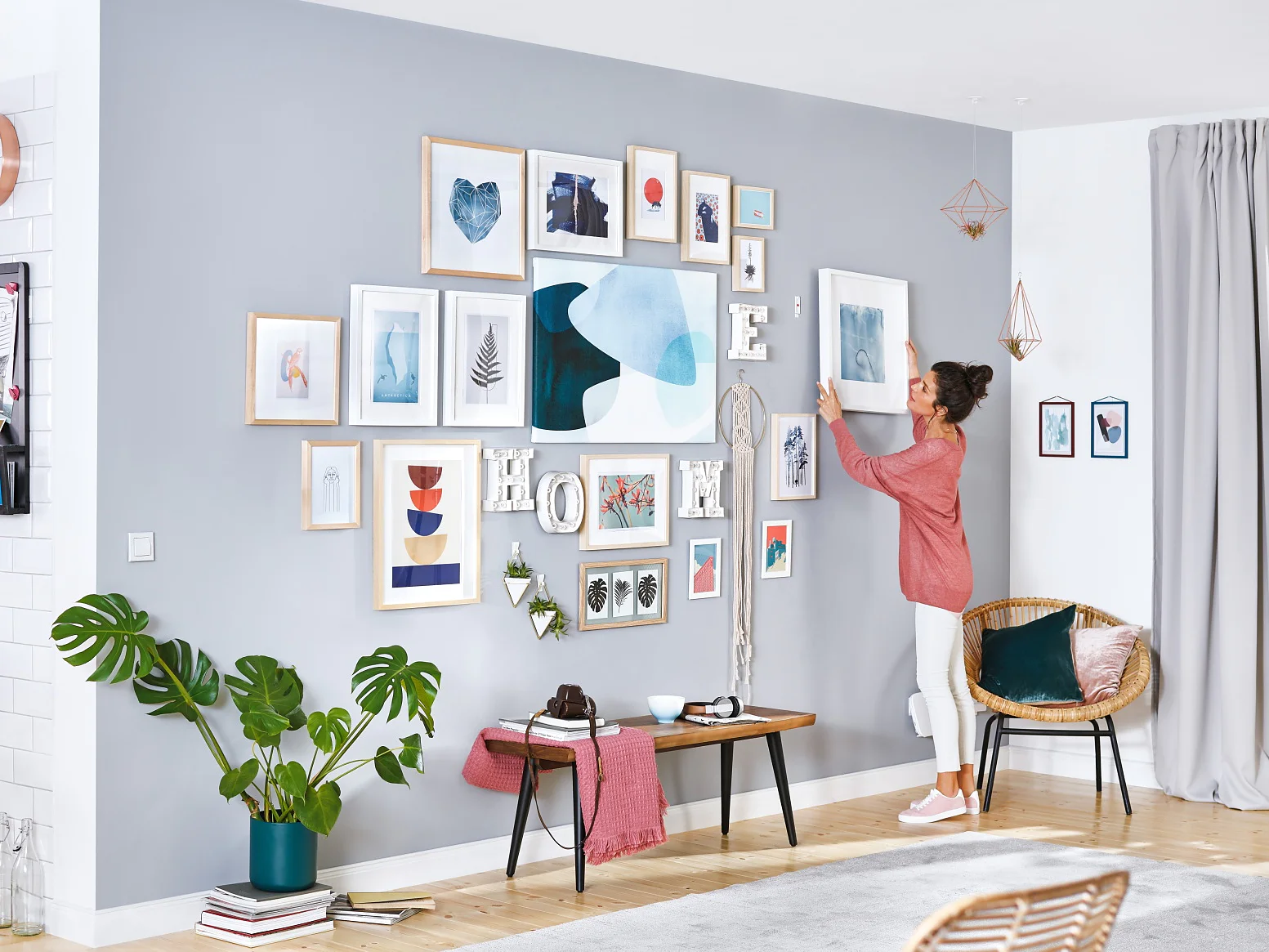 A woman decorates a living room wall with various framed artworks, using tesa tape. The room features a light gray wall, wooden flooring, and modern furniture, including a wooden table and chairs. A kitchen is visible in the background. (This text has been generated by AI)