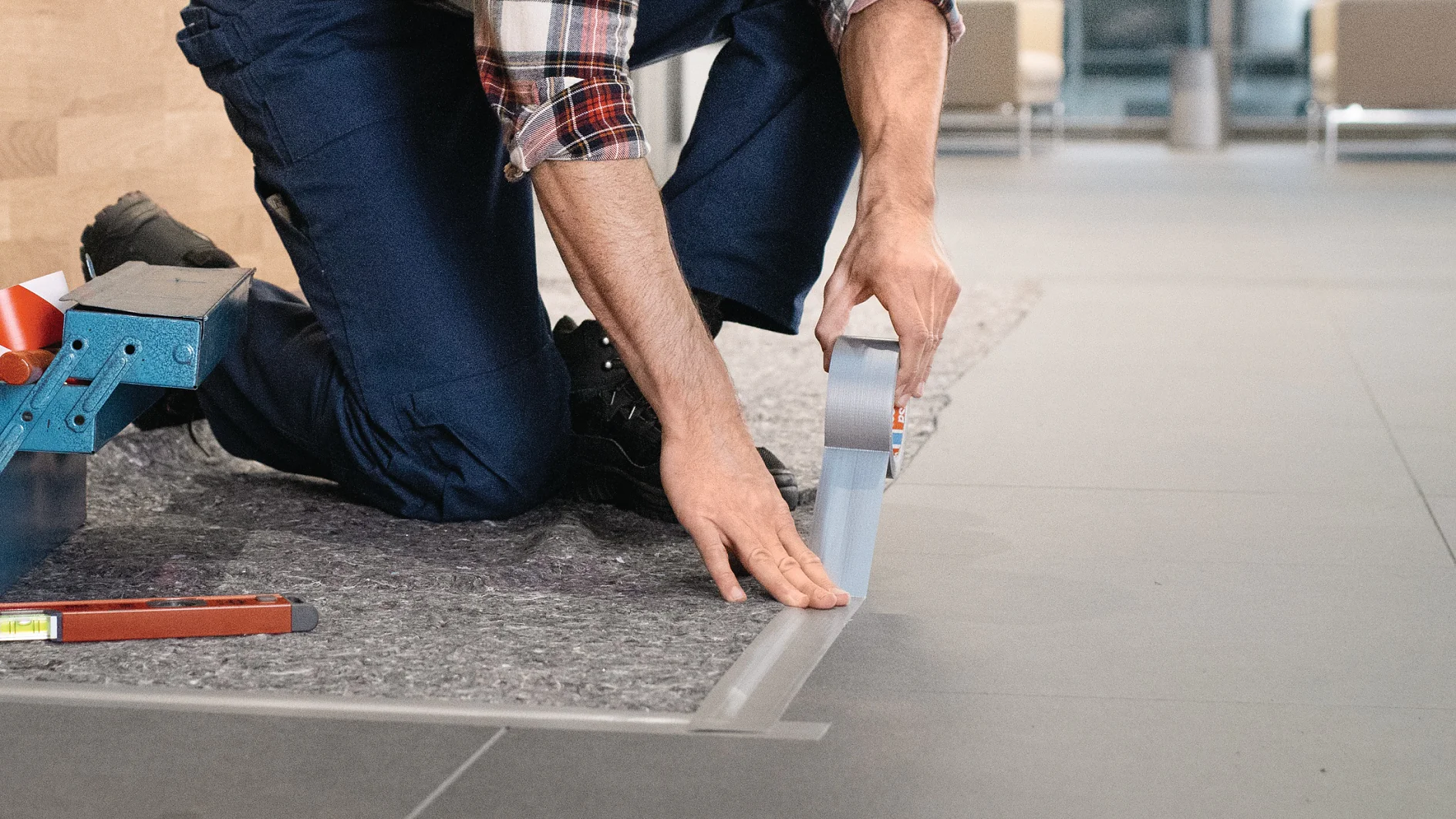 A person kneels on the floor, applying silver tesa tape to the edge of a gray carpet on a tiled surface. They are wearing a plaid shirt and blue pants. A toolbox with tools is visible to the left. (This text has been generated by AI)