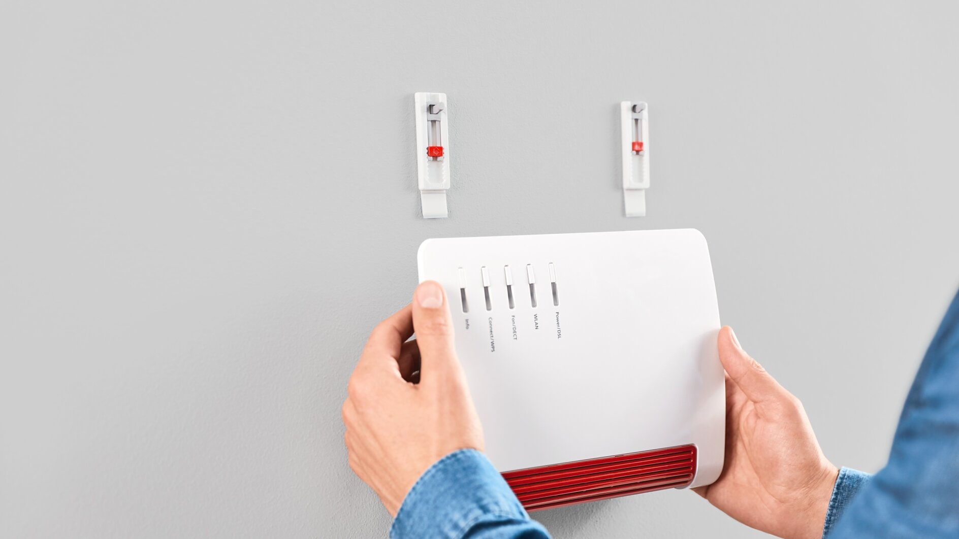 Osoba w niebieskiej koszuli montuje białe urządzenie elektroniczne z czerwonymi akcentami na szarej ścianie. Ściana ma dwa haki, a osoba wyrównuje urządzenie, które ma etykiety ze wskaźnikami i prawdopodobnie jest routerem lub modemem. Używa taśmy tesa, aby bezpiecznie przymocować urządzenie do ściany. (This text has been generated by AI)