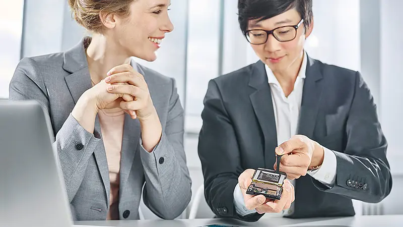 Two business professionals in suits discussing a smartphone at a desk with documents and a laptop in a bright office. (This text has been generated by AI)