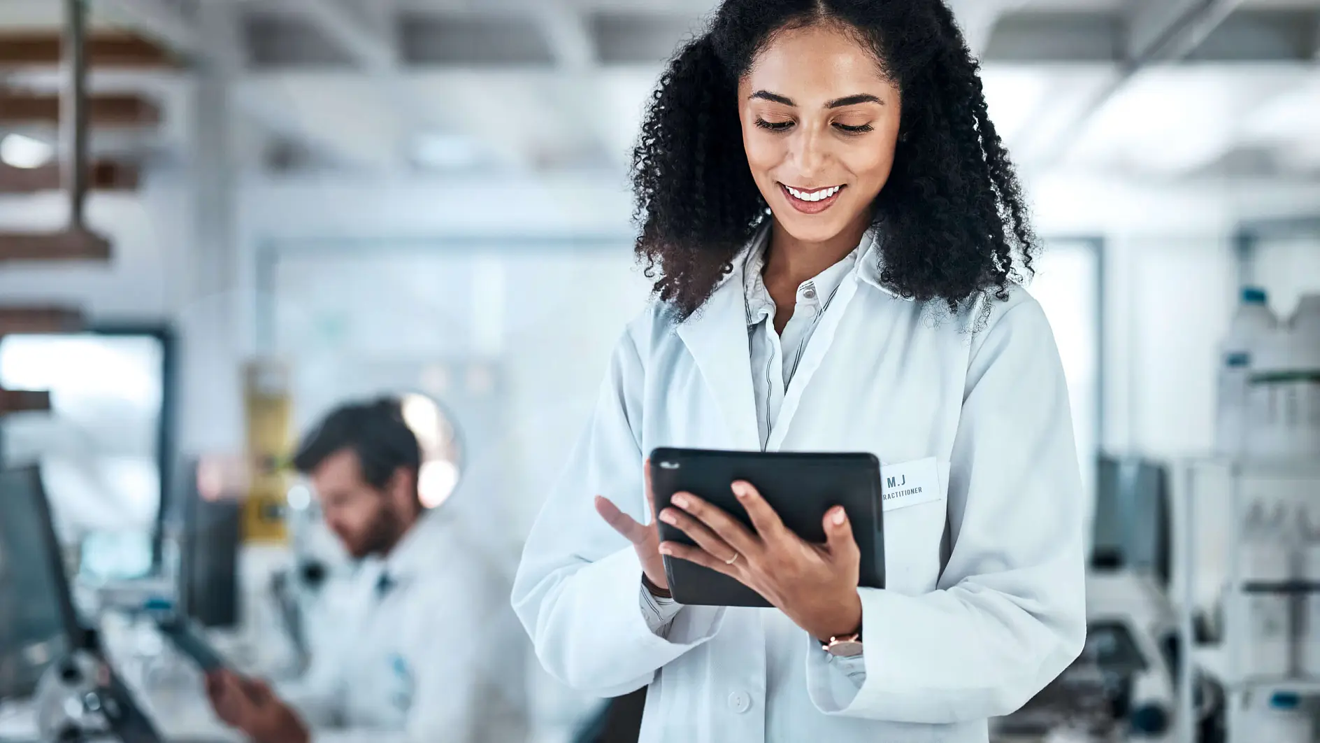 scientist in white lab coat using tablet in modern laboratory with colleague working in background
