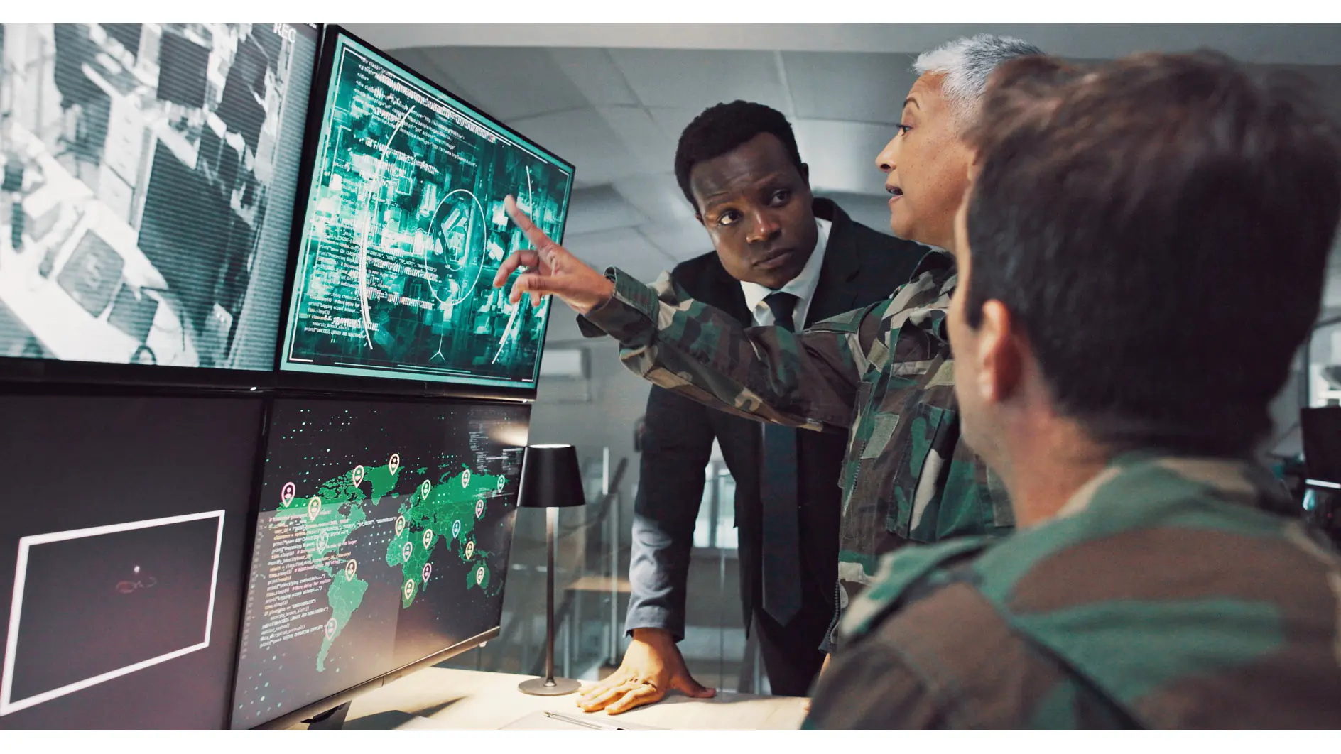 Trzy osoby w ubraniach wojskowych i biznesowych analizują cyfrowe mapy na monitorach w centrum kontroli; jedna z nich wskazuje dane za pomocą taśmy tesa. (This text has been generated by AI)