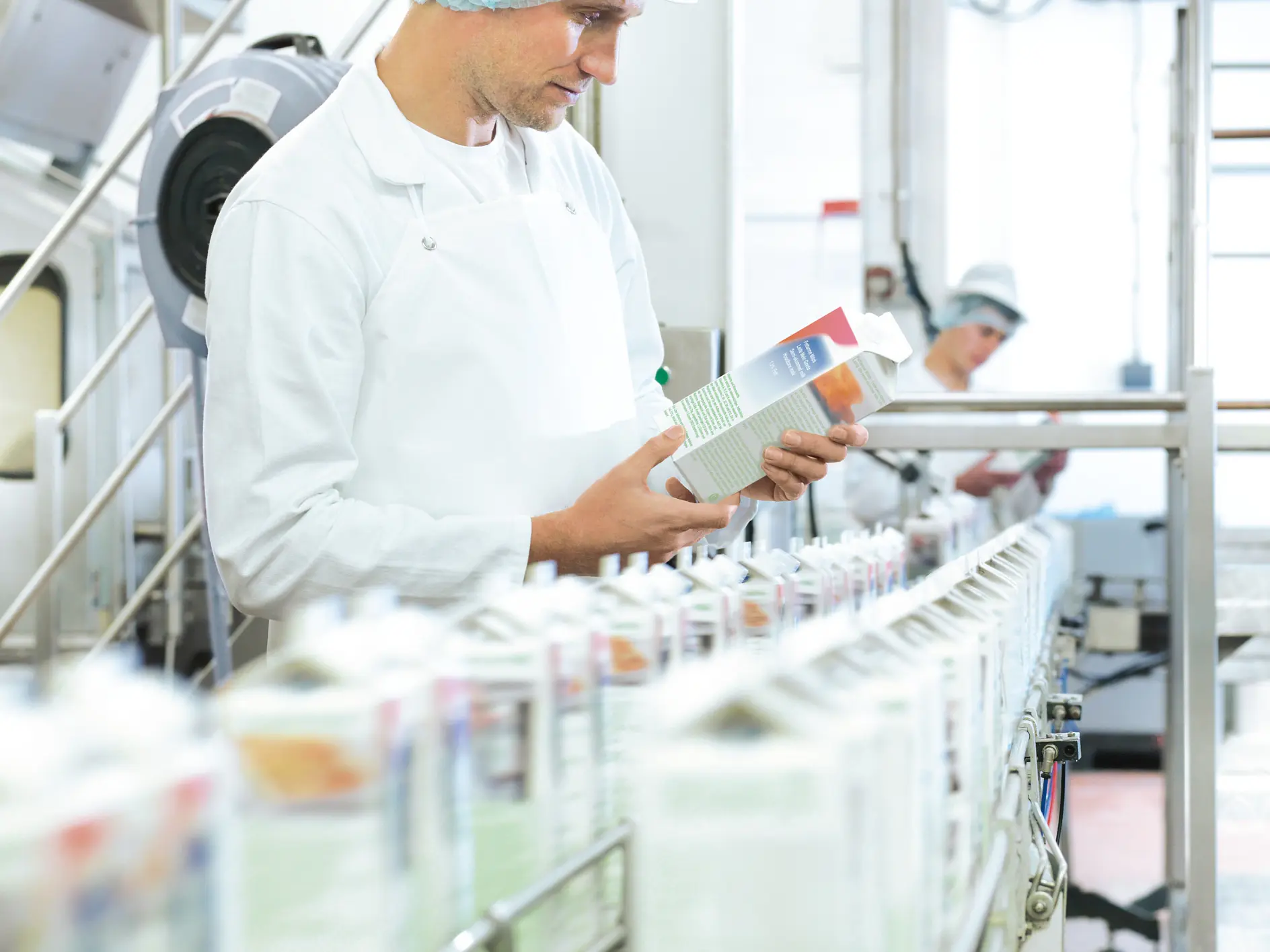 Um operário de fábrica usando avental branco, touca e gorro inspeciona uma caixa em uma linha de montagem cheia de caixas semelhantes, seladas com fita tesa. Outro trabalhador ao fundo também veste roupas similares em um ambiente industrial bem iluminado. (This text has been generated by AI)