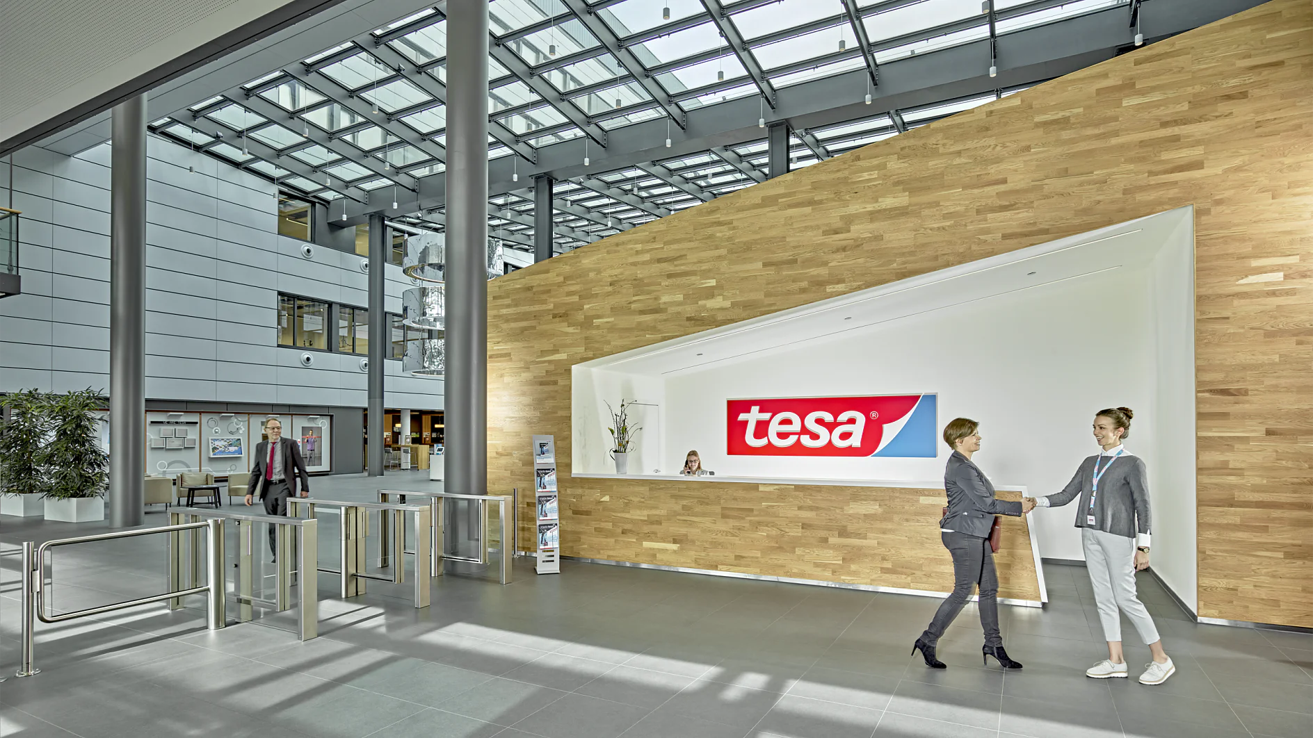 Um lobby de escritório moderno com uma mesa de recepção exibindo o logotipo da tesa. Duas pessoas estão apertando as mãos em frente à mesa. O espaço tem paredes de vidro, teto alto e catracas. Uma pessoa está caminhando ao fundo. (This text has been generated by AI)