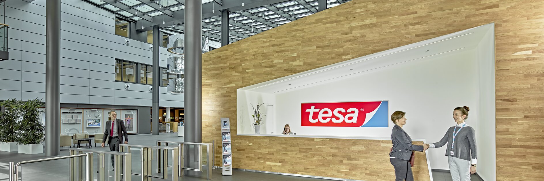Um lobby de escritório moderno com uma mesa de recepção exibindo o logotipo da tesa. Duas pessoas estão apertando as mãos em frente à mesa. O espaço tem paredes de vidro, teto alto e catracas. Uma pessoa está caminhando ao fundo. (This text has been generated by AI)