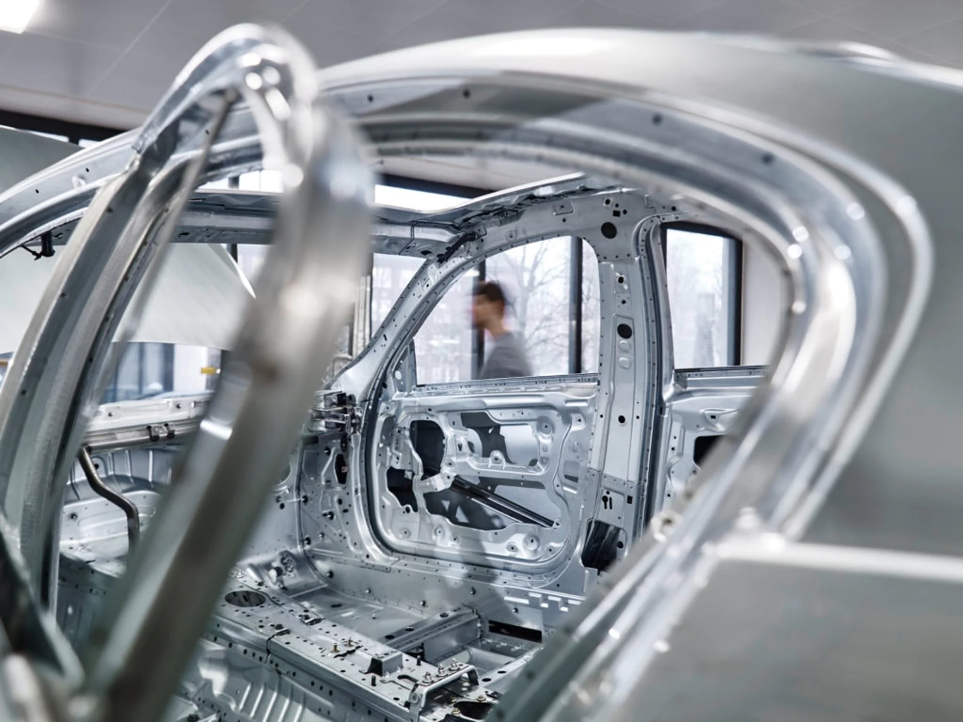 Fechamento de furos na produção Uma estrutura de carro parcialmente montada com estruturas metálicas visíveis é mostrada em um ambiente industrial. O fundo apresenta grandes janelas com vista para uma cena ao ar livre. Uma figura borrada é visível à distância e, perto, a fixação confiável da fita tesa prende os componentes de forma eficaz. (This text has been generated by AI)