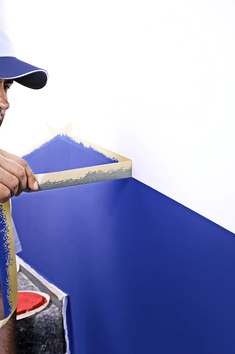 A man wearing a blue cap and a white shirt with blue overalls is painting a geometric design on a wall. He is peeling tesa tape from a blue-painted section on a white wall. A paint can is visible at his side. (This text has been generated by AI)