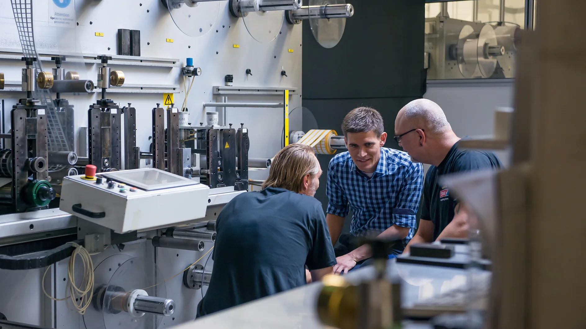 Three men are gathered around a large industrial machine in a factory setting. They appear to be engaged in discussion. The machine has various levers and components, and there are rolls of tesa tape in the background. (This text has been generated by AI)