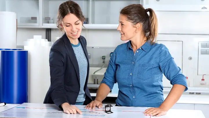 Two women in a laboratory setting discuss a project over a large sheet of paper on a table. One woman points to the paper while the other listens attentively. The room has lab equipment and storage cabinets in the background, and they use tesa tape to secure various documents and samples in place as part of their work.