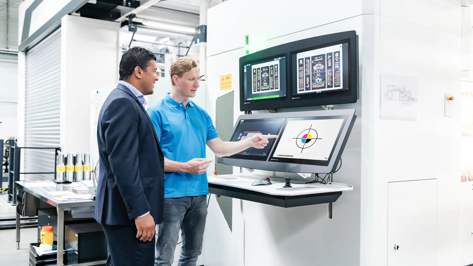 Two men stand in front of a control panel with multiple screens in an industrial setting. One wears a suit, the other a blue polo shirt and jeans. The man in the polo points at a screen displaying technical graphics. Equipment is visible in the background, including rolls of tesa tape prominently displayed nearby.