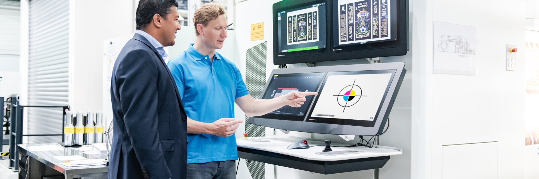 Two men stand in front of a control panel with multiple screens in an industrial setting. One wears a suit, the other a blue polo shirt and jeans. The man in the polo points at a screen displaying technical graphics. Equipment is visible in the background, including rolls of tesa tape prominently displayed nearby.