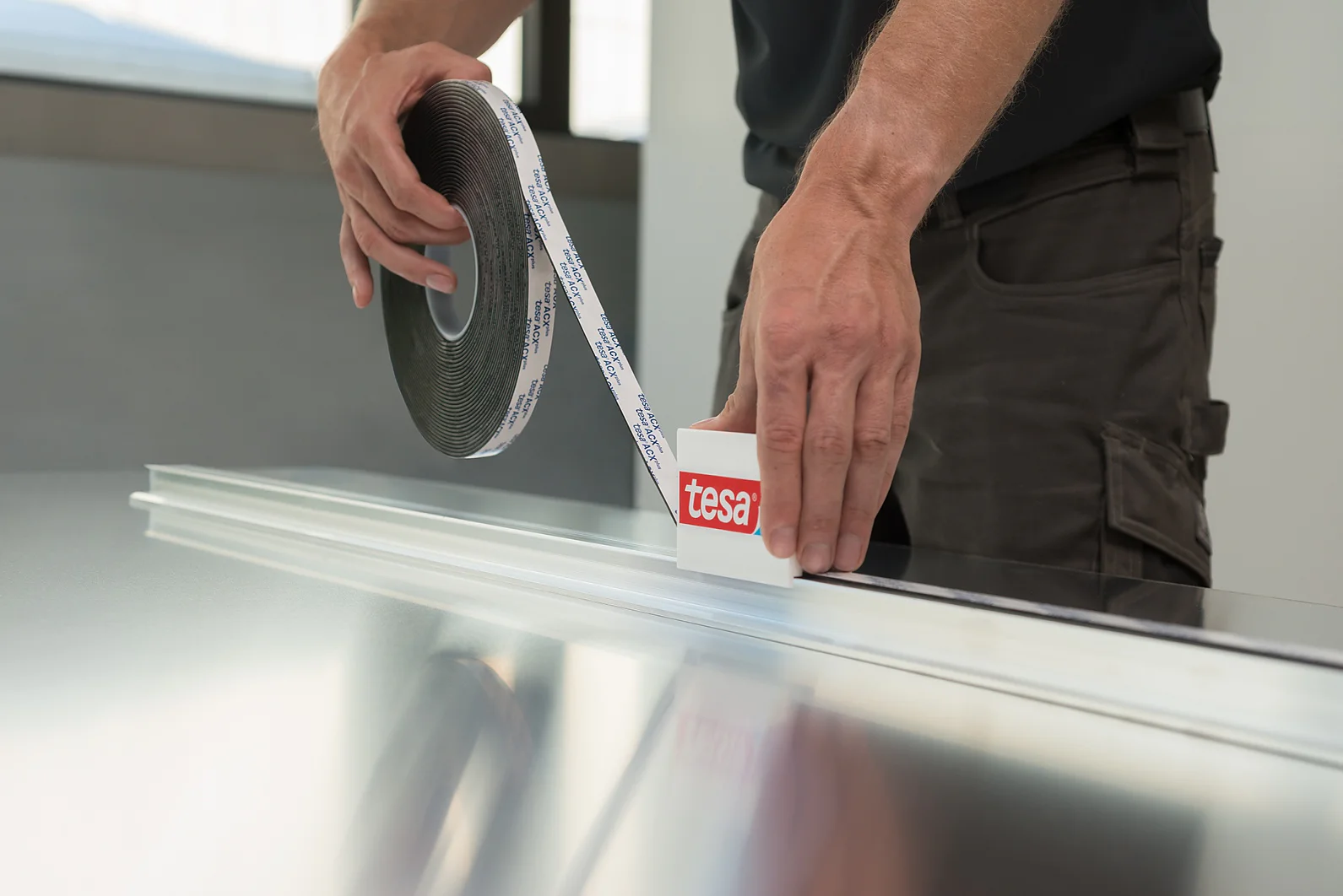A person applies tesa tape from a roll onto a long, flat surface. The tesa tape roll has a label with the word tesa. The person is wearing a dark shirt and brown pants, and the surface appears to be metallic. (This text has been generated by AI)