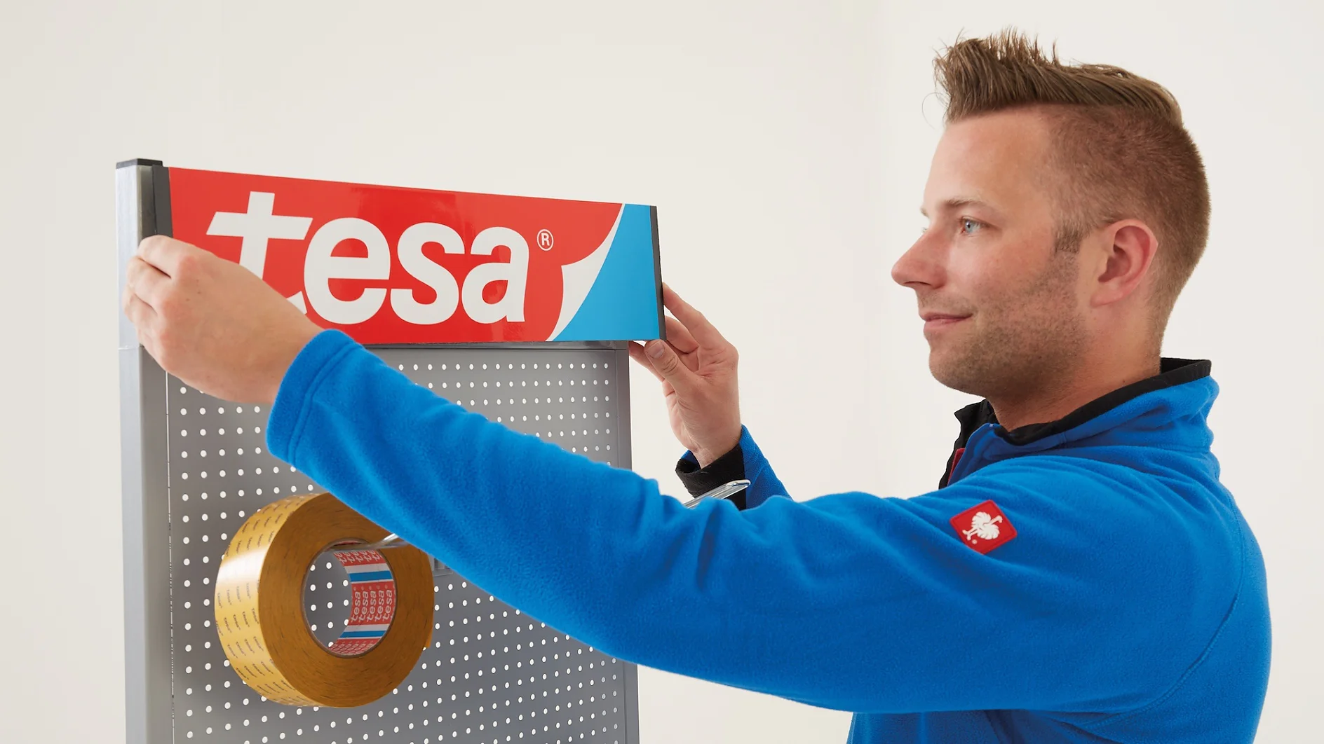 Uma pessoa de jaqueta azul está ajustando um painel de exposição com uma grande placa vermelha e branca tesa. Um rolo de fita tesa está pendurado no painel perfurado. O fundo é branco liso. (This text has been generated by AI)