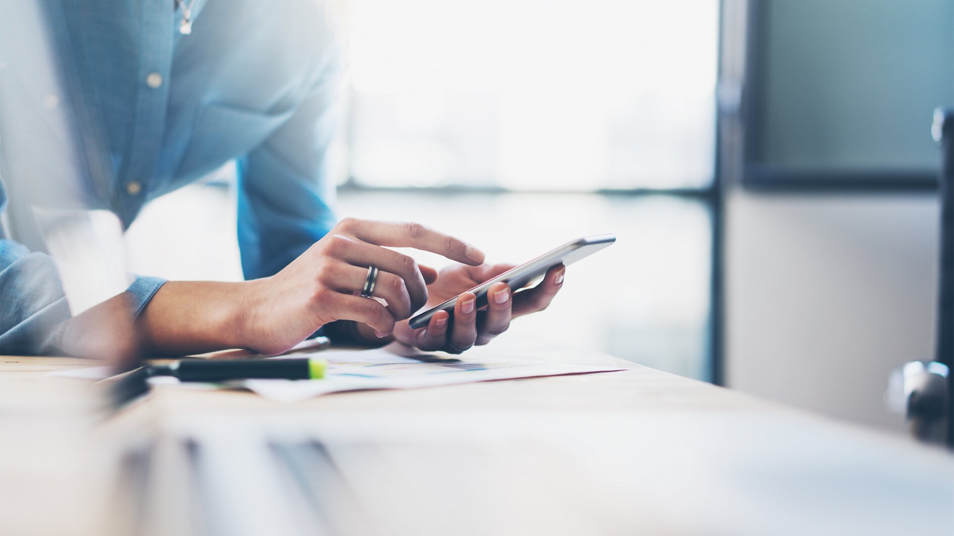 A person in a blue shirt is standing at a desk, using a smartphone. The background is blurred. Papers and a laptop are on the desk in the foreground, along with tesa tape. Sunlight is streaming in through a window. (This text has been generated by AI)