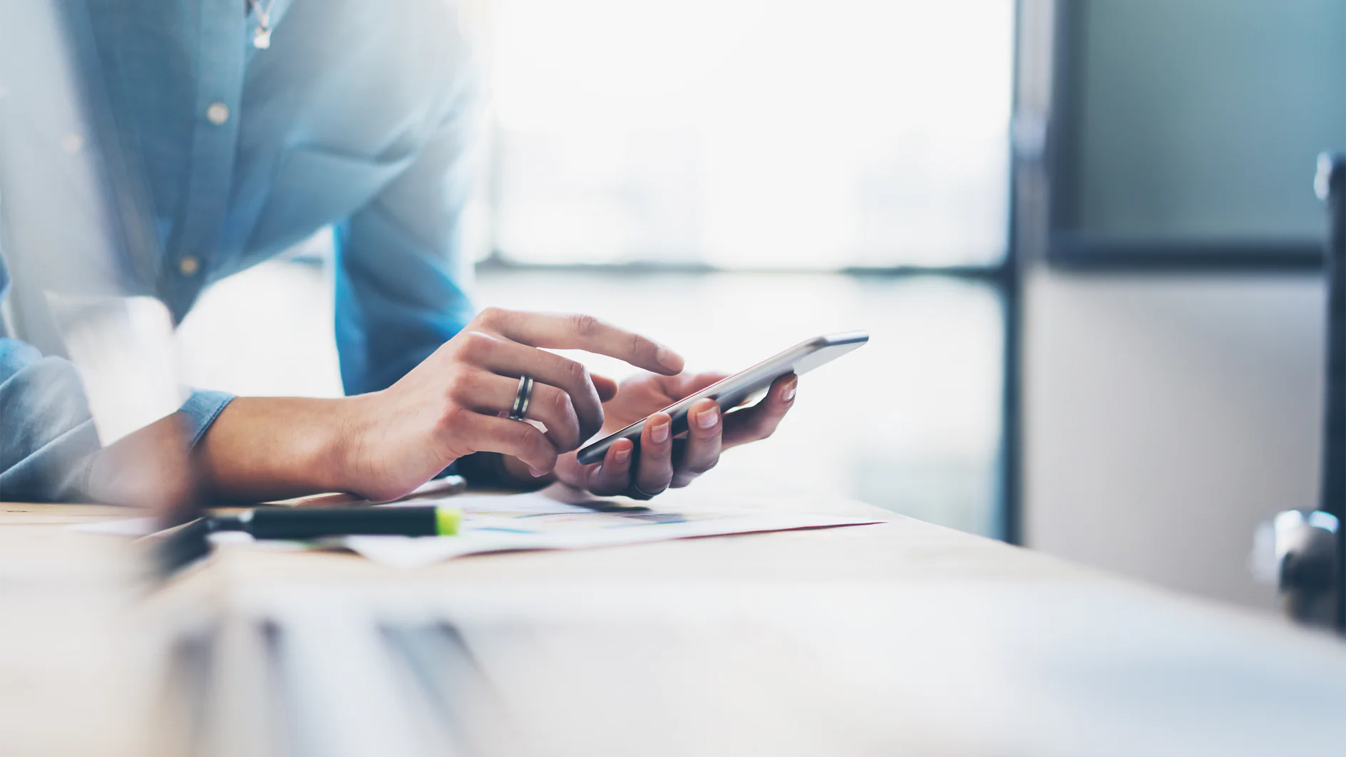 A person in a blue shirt is standing at a desk, using a smartphone. The background is blurred. Papers and a laptop are on the desk in the foreground, along with tesa tape. Sunlight is streaming in through a window. (This text has been generated by AI)