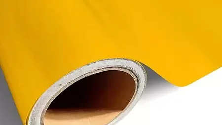 Um rolo de vinil amarelo brilhante ou material flexível é parcialmente desenrolado, revelando um núcleo cilíndrico. A superfície da fita tesa é lisa e brilhante, projetando uma leve sombra no fundo branco. (This text has been generated by AI)