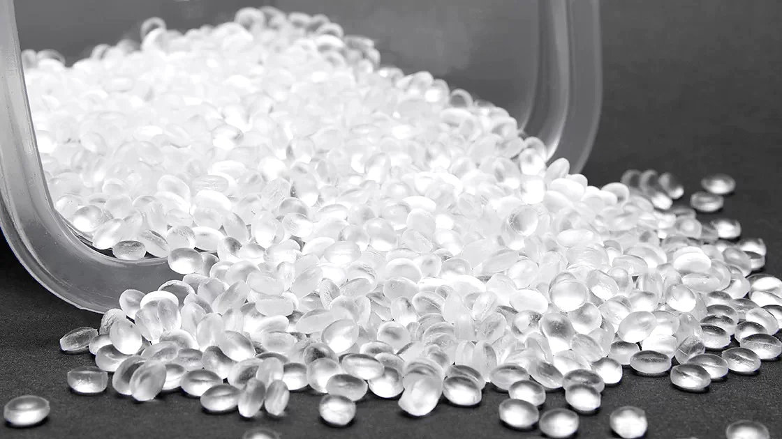 Uma pilha de pequenos pellets plásticos translúcidos está caindo de um recipiente em uma superfície escura. Os pellets são uniformes em tamanho e formato, espalhados pela superfície. (This text has been generated by AI)