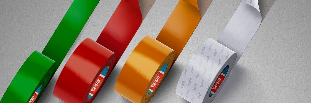Quatro rolos de fita tesa são exibidos verticalmente, apresentando as cores verde, vermelho, amarelo e branco. As fitas tesa são parcialmente desenroladas com uma etiqueta de marca visível dentro de cada rolo. O fundo é um cinza neutro. (This text has been generated by AI)