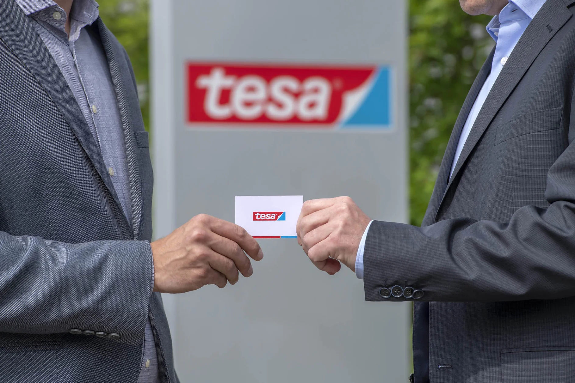 Dois indivíduos em trajes de negócios trocam um cartão com um logotipo vermelho e azul de tesa tape. Uma placa maior com o mesmo logotipo é visível ao fundo. O cenário parece ser ao ar livre. (This text has been generated by AI)