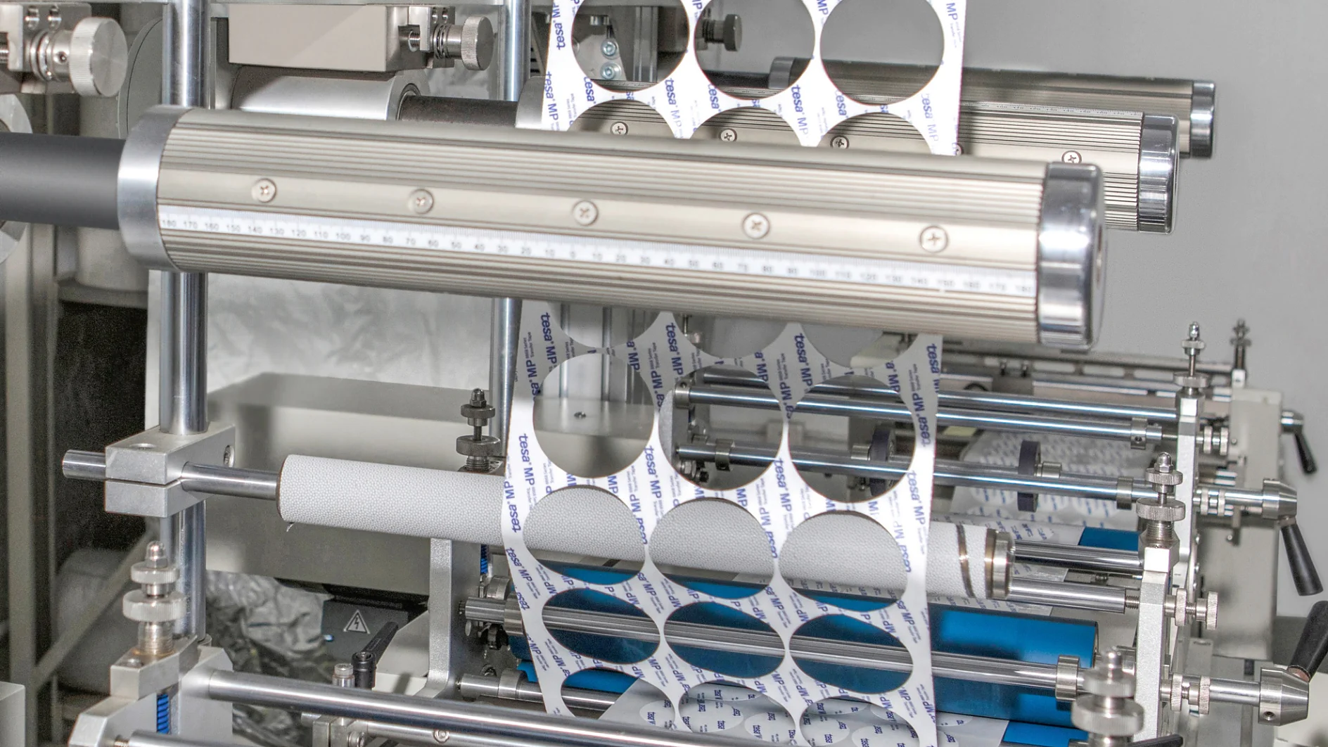 Máquinas industriais imprimindo e cortando etiquetas adesivas em um rolo de fita tesa. A máquina exibe fileiras de etiquetas circulares, algumas das quais são cortadas. O cenário parece ser um ambiente de fabricação. (This text has been generated by AI)