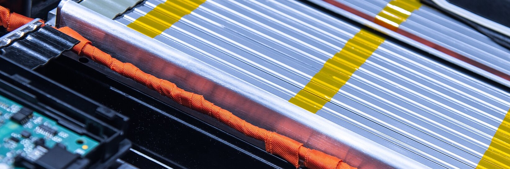 Close-up of a complex electrical circuit board and battery pack, featuring silver and yellow striped battery cells, orange wires, and a green circuit board. The components are tightly arranged within a metallic frame using tesa tape. (This text has been generated by AI)